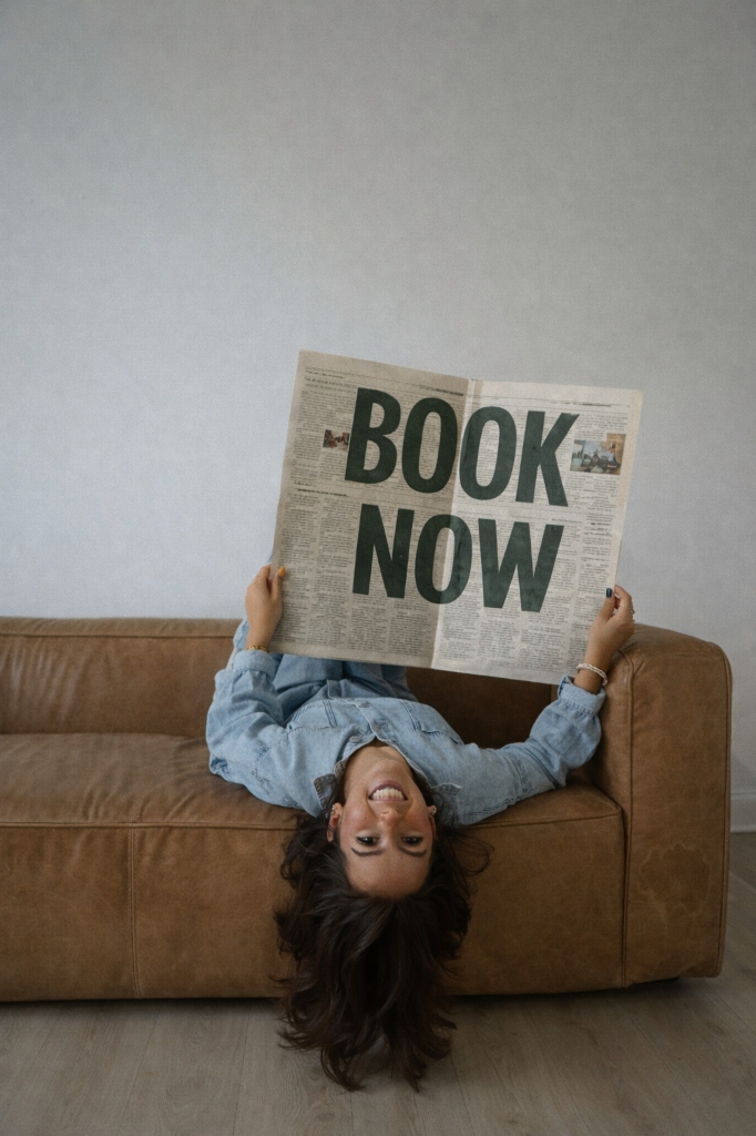 Woman upside down on sofa holding Book Now newspaper
